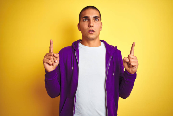 Young handsome man wearing purple sweatshirt over yellow isolated background amazed and surprised looking up and pointing with fingers and raised arms. - Foto, Imagen