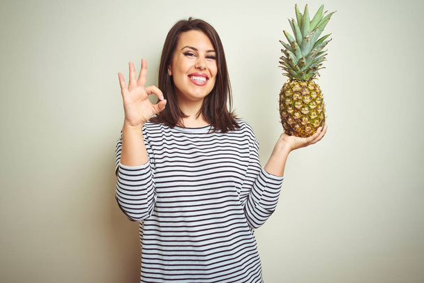 Young beautiful woman holding tropical fruit pineapple over isolated background doing ok sign with fingers, excellent symbol - Foto, Bild