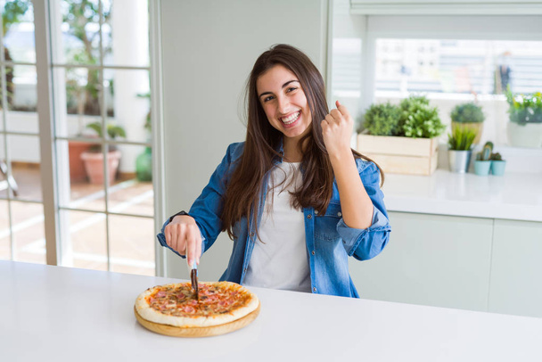 Mulher bonita cortando uma fatia de pizza saborosa usando um cortador gritando orgulhoso e celebrando vitória e sucesso muito animado, torcendo emoção
 - Foto, Imagem