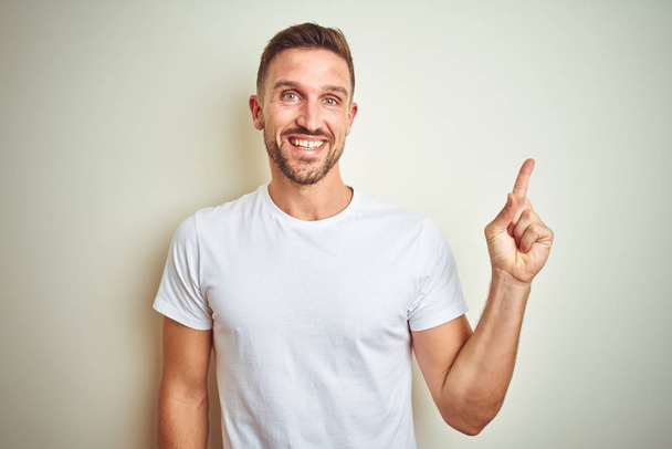 Mladý pohledný muž nosí ležérní bílé tričko izolované pozadí s velkým úsměvem na tváři, rukou a prstu přejdete na straně při pohledu na fotoaparát. - Fotografie, Obrázek
