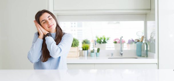 Wide angle picture of beautiful young woman sitting on white table at home sleeping tired dreaming and posing with hands together while smiling with closed eyes. - Photo, Image