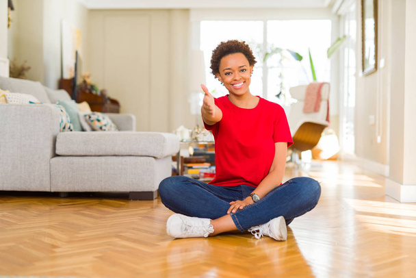 Nuori kaunis afrikkalainen amerikkalainen nainen istuu lattialla kotona hymyillen ystävällinen tarjoamalla kädenpuristus tervehdys ja kutsuva. Menestyksekäs yritys
. - Valokuva, kuva