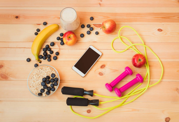 Glass of yogurt, fruits, oat flakes, jumping rope, dumbbells and smartphone on wooden background.  - Foto, imagen
