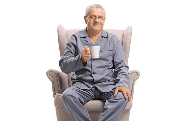 Elderly man in pajamas sitting in an armchair and holding a cup of hot drink isolated on white background - 写真・画像
