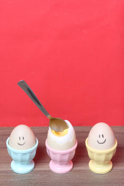 A boiled egg with a chopped shell stands in a poached. On the egg painted smiley. A spoon is stuck in it for food. Against the background of coral color. - Foto, Bild