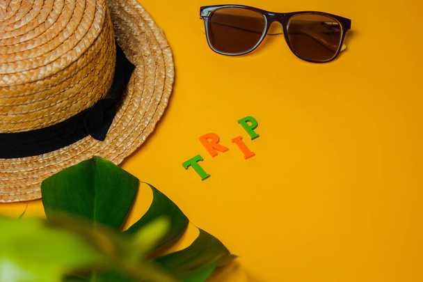 Beach accessories: straw hat, sunglasses, the inscription trip and a wide leaf of a monstera plant for shade on a bright yellow background on a sunny day, top view, copy space - 写真・画像
