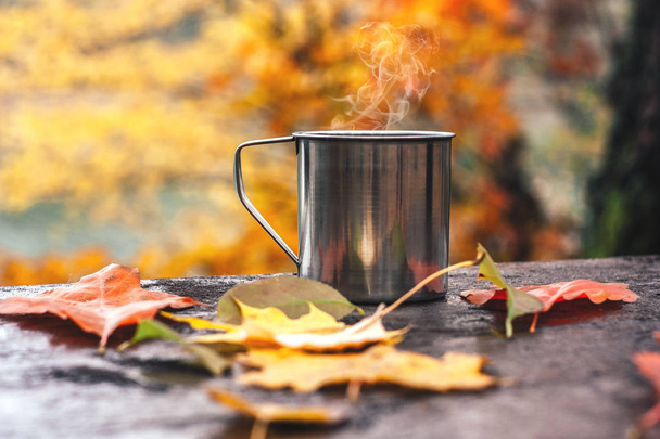 Hot drink in the steel cup on the wooden table. Autumn orange leaves. - Photo, Image