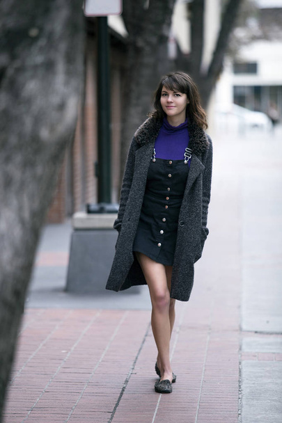 Young pretty woman walking on an urban street sidewalk or a pavement city park.  She looks like an independent millennial female tourist wearing a coat traveling solo on foot.  - Photo, Image