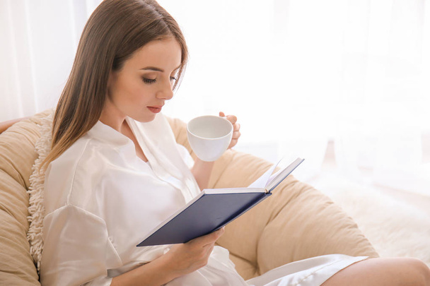 Beautiful young woman drinking coffee while reading book at home - Фото, изображение