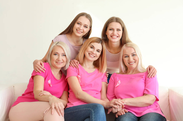 Όμορφες γυναίκες διαφορετικών ηλικιών με ροζ κορδέλες που κάθονται στον καναπέ στο δωμάτιο. Έννοια του καρκίνου του μαστού - Φωτογραφία, εικόνα