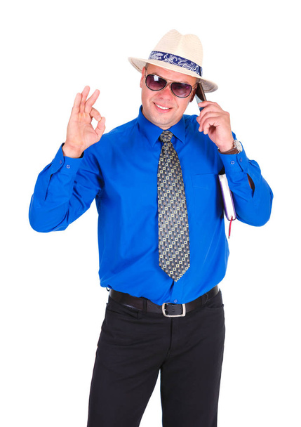 Счастливый и успешный бизнесмен в синей рубашке, галстуке, солнцезащитных очках и белой шляпе. Говорит по мобильному телефону, держит блокнот под мышкой и показывает жест Хорошо. Изолированный белый фон
 - Фото, изображение