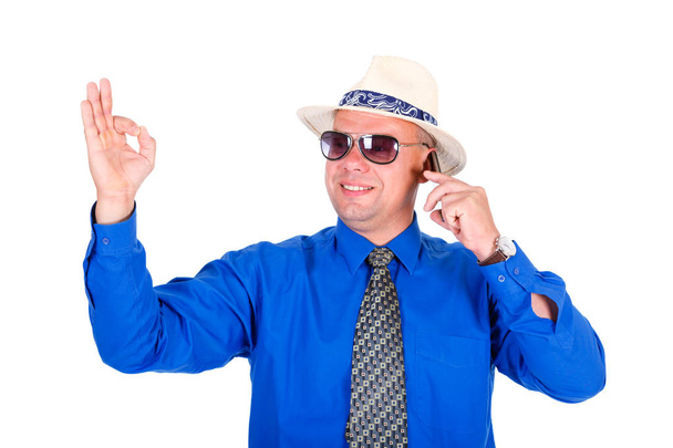 Успешный и улыбающийся бизнесмен в синей рубашке, галстуке, солнцезащитных очках и белой шляпе. Выступление на мобильном телефоне и показывает жест ОК. Изолированный белый фон, концепция лидерства и успеха
 - Фото, изображение