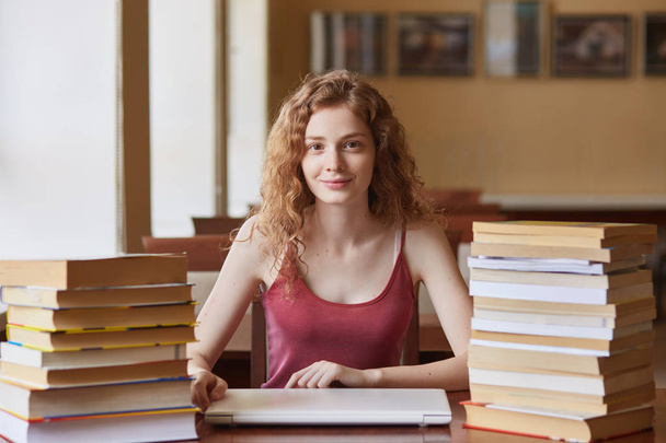 Shot of young foxy haired female student sitting at table in library, preparing for test or exam, posing with pleasant facial expression, looking concentrated dierectly at camera. Education concept. - Photo, image