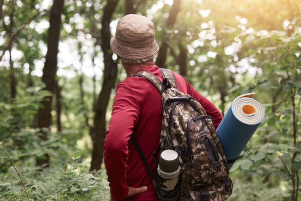 Outdoor shot of old man having bag with thermos and sleeping pad, wearing beige hat and red sweatshirt, looking for adventures in forest alone, being fond of traveling and hiking. Hobby concept. - Foto, Bild