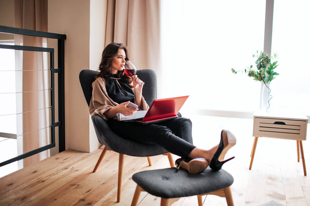 Young businesswoman work at home. Drinking red wine from glass and look to right. Hold phone in hands. Journal and laptop on legs. Remote work. Alone in room. Relaxing. - Foto, imagen