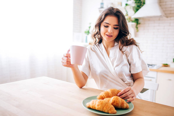 Young woman with dark hair Sitting in kitchen and drinking coffee in morning. Taking one croissant and smile. Delightful model. - Foto, immagini