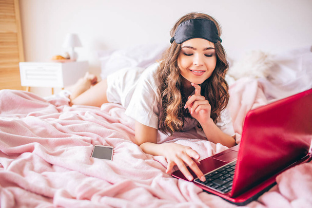 Young woman with dark hair lying on bed in bedroom. Using laptop and look at it. Cheerful positive model typing on keybaord. Cellphone on bed. - Fotoğraf, Görsel