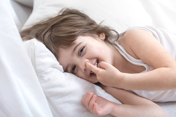 Χαριτωμένο κοριτσάκι χαμογελά ενώ βρίσκεται σε ένα άνετο λευκό κρεβάτι - Φωτογραφία, εικόνα