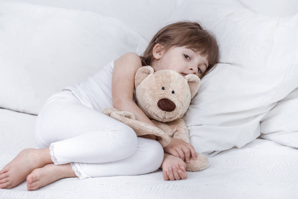 Мила дівчинка посміхається, лежачи в затишному білому ліжку
 - Фото, зображення