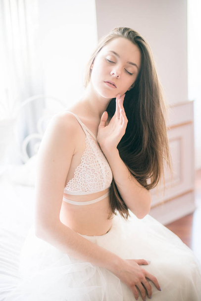 Young beautiful bride with dark long hair morning at home on a white bed in a white lace bustier and dress - Foto, Imagen