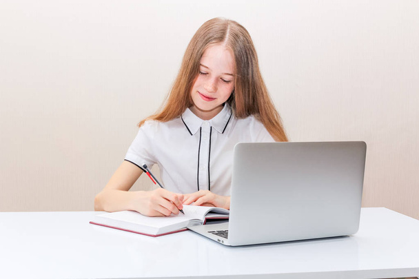 junges Mädchen mit blonden Haaren in weißer Bluse sitzt am Tisch und blickt in den Laptop-Bildschirm - Foto, Bild