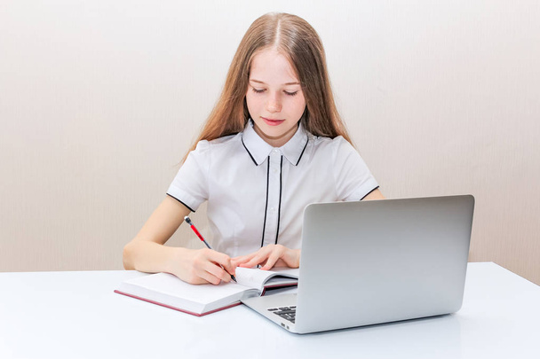 junges Mädchen mit blonden Haaren in weißer Bluse sitzt am Tisch und blickt in den Laptop-Bildschirm - Foto, Bild