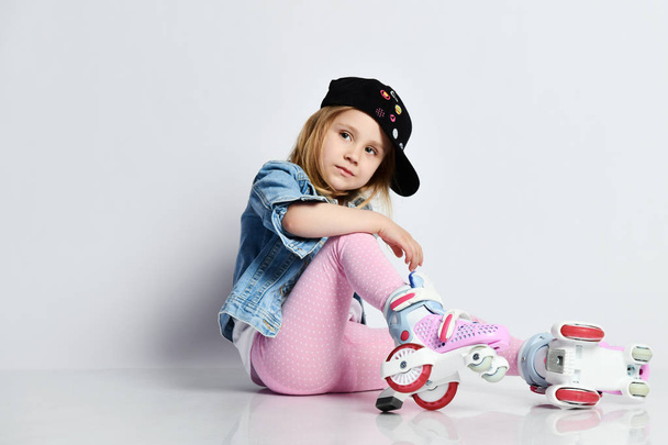 girl sits on the rollers in a blue denim and cap and smiles. Summer sports for children. - Фото, изображение