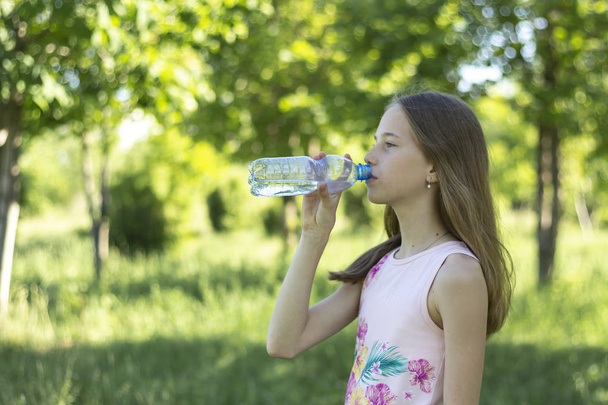 Een jong blond meisje in een roze t-shirt drinkt water uit een plastic fles op een achtergrond van groen bos - Foto, afbeelding