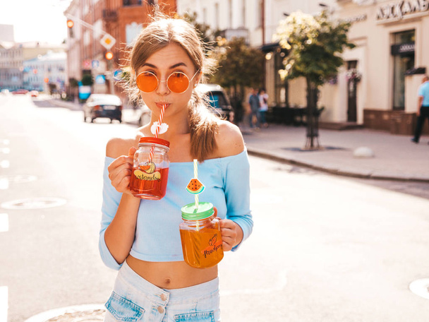 Young beautiful smiling hipster woman in sunglasses.Girl in summer trendy clothes posing in the street.Model holding and drinking two fresh cocktail smoozy drink - Foto, Bild