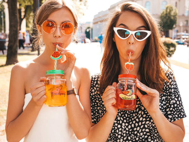 Two young beautiful smiling hipster girls in trendy summer clothes,dress.Sexy carefree women posing in the street.Positive models having fun in sunglasses.Drinking fresh cocktail smoozy drink - Fotó, kép