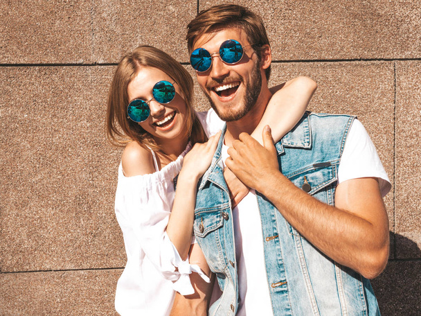 Smiling beautiful girl and her handsome boyfriend. Woman in casual summer dress.Man in jeans clothes.Happy cheerful family.Female having fun on the street near wall.Hugging couple in sunglasses - 写真・画像