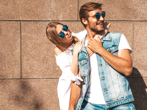 Smiling beautiful girl and her handsome boyfriend. Woman in casual summer dress.Man in jeans clothes.Happy cheerful family.Female having fun on the street near wall.Hugging couple in sunglasses - Foto, afbeelding