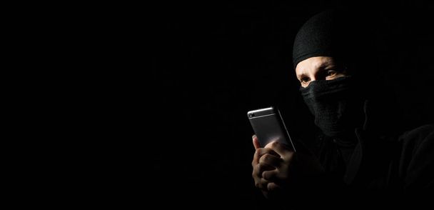 Close-up Of Hacker Using Smart Phone. Young adult girl in black clothes with hidden face looks at smartphone screen on black background with copy space. - Valokuva, kuva