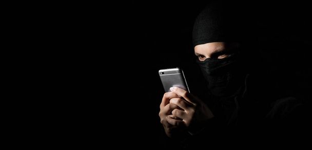 Close-up Of Hacker Using Smart Phone. Young adult girl in black clothes with hidden face looks at smartphone screen on black background with copy space. - Foto, Imagem
