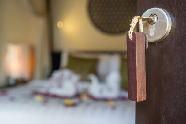 Vista de perto na chave do quarto de hotel na fechadura, interior do quarto do hotel borrado com cisnes de toalha e flores de rosa na cama
 - Foto, Imagem