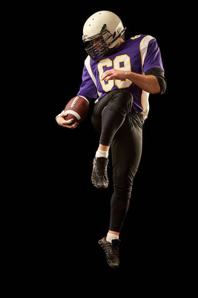 American football player holding a ball while jumping away from a strike. Black background, copy space. Violet with white sportswear. - Photo, image