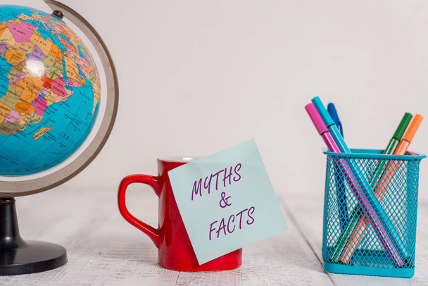 Writing note showing Myths And Facts. Business photo showcasing usually traditional story of ostensibly historical events Globe map world earth coffee cup sticky note pens holder wooden table. - Photo, Image