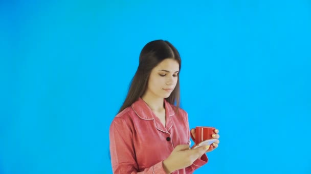 Happy girl in pajamas is drinking tea or coffee and using phone on blue background in studio - Кадри, відео