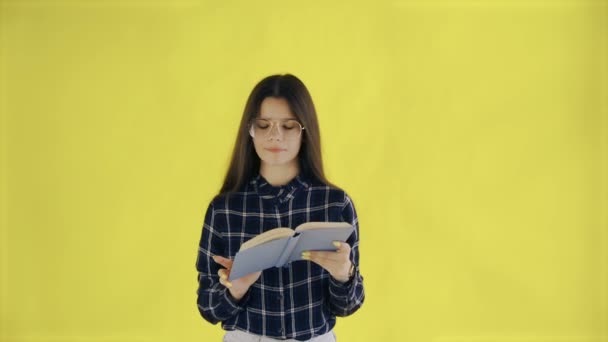 Beautifu young girl in glasses is reading book isolated on yellow background - Záběry, video