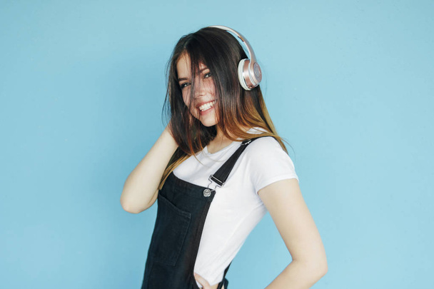 Счастливая беззаботная улыбающаяся девочка-подросток с темными длинными волосами в белой футболке, слушающая музыку в наушниках, изолированных на синем фоне
 - Фото, изображение