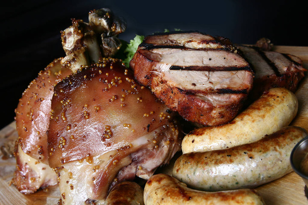 Assorted grilled meat. Knuckle, steaks, chicken (duck)? potatoes on a wooden board. Close up on a black background. - Photo, Image