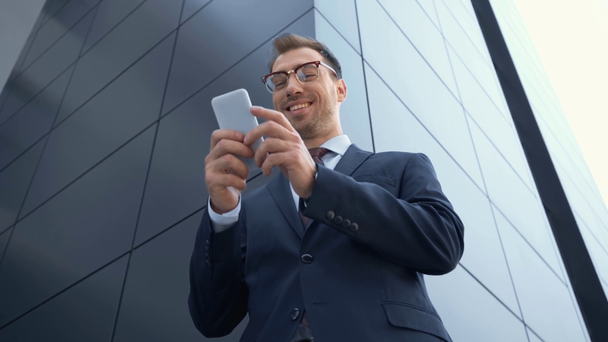 visão de baixo ângulo de homem de negócios usando smartphone e sorrindo perto do edifício
 - Filmagem, Vídeo