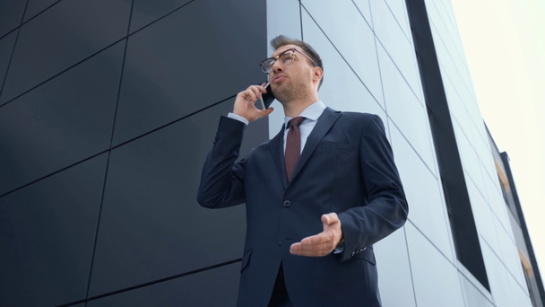 vista de bajo ángulo del hombre de negocios utilizando el teléfono inteligente, gestos y sonriendo cerca del edificio
 - Metraje, vídeo