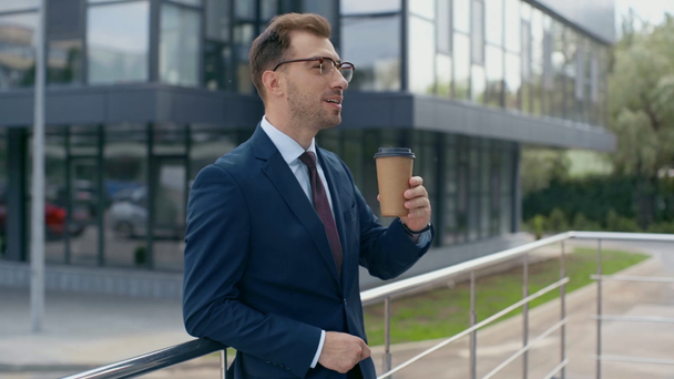 selective focus of businessman drinking coffee to go, smiling and looking at camera near building - Footage, Video