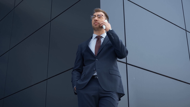 Низкоугольный вид бизнесмена в формальной одежде, улыбающегося и говорящего по смартфону возле здания
 - Кадры, видео