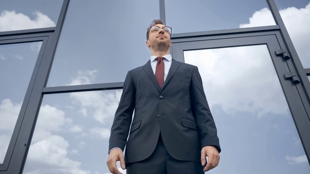 Niederwinkelblick des Geschäftsmannes, der sich umschaut, die Hand hebt und auf die Uhr in der Nähe des Gebäudes blickt - Filmmaterial, Video