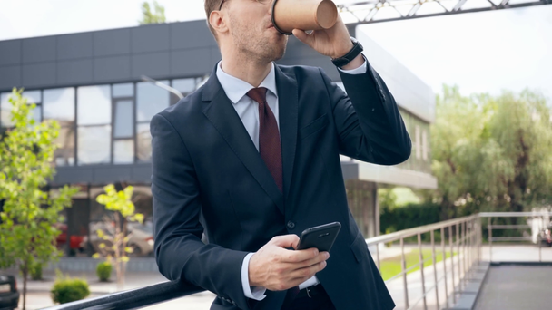 businessman using smartphone, smiling and drinking coffee to go near building - Footage, Video
