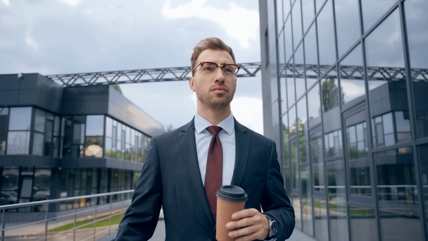 眼鏡をかけたビジネスマンとフォーマルな服装で、建物の近くに行くためにコーヒーを歩いたり飲んだりする - 映像、動画