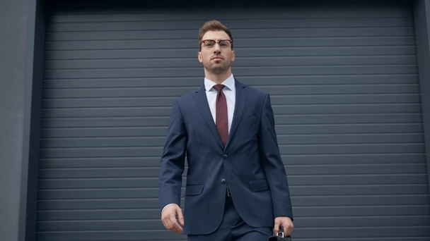 灰色の建物の近くを歩くブリーフケースを持つ自信のあるビジネスマン - 映像、動画