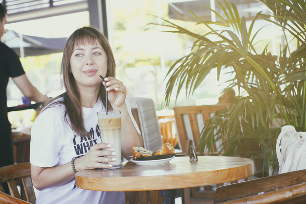 Młoda dziewczyna siedzi w kawiarni z kieliszek zimnej kawy z mlekiem i ciesząc słoneczny letni dzień.  - Zdjęcie, obraz